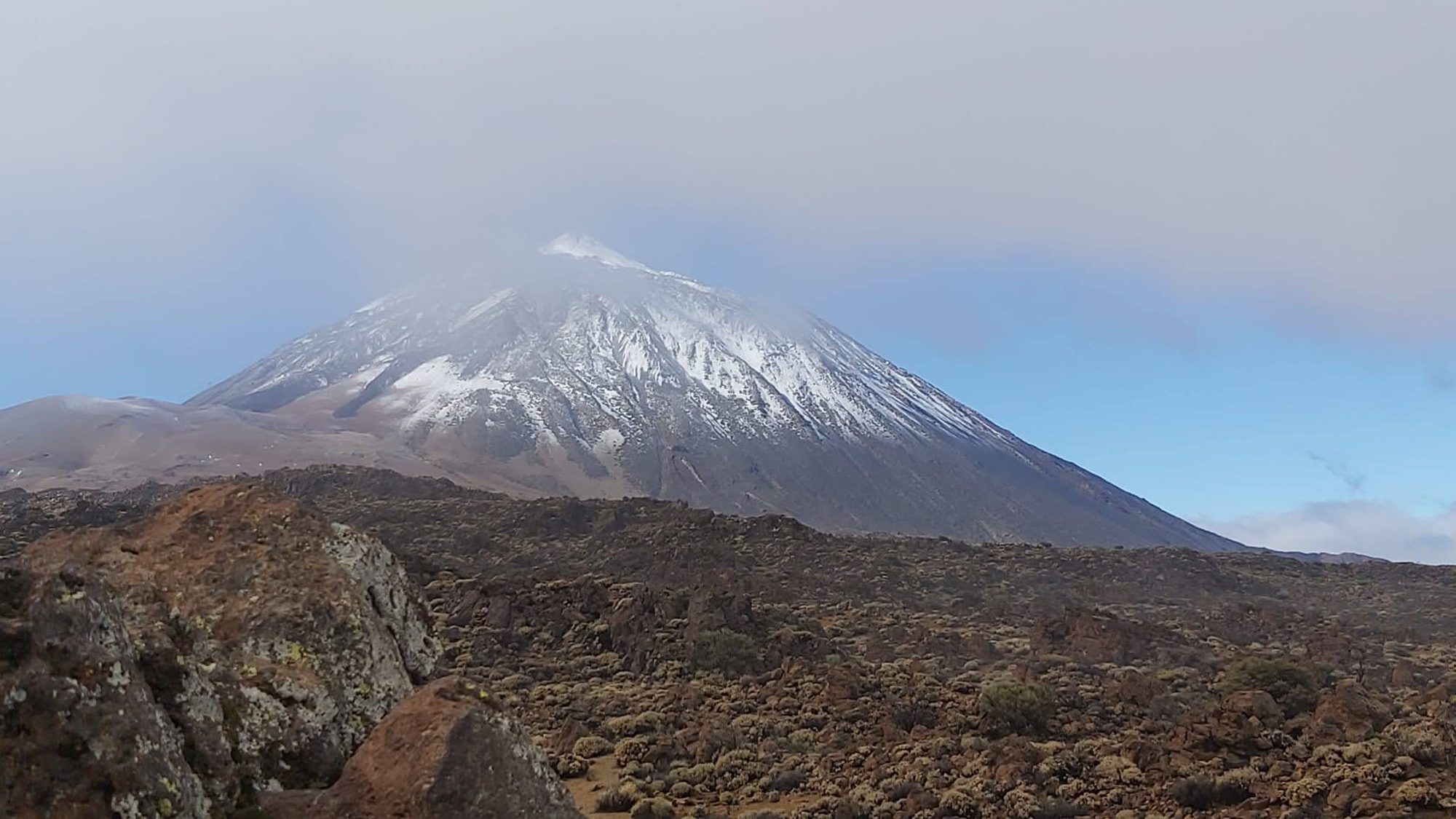 Pico del Teide nevado sobre un paisaje volcánico y rocoso bajo cielo nublado.